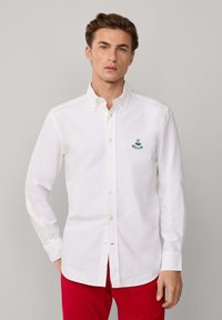 Hombre vestido con una camisa blanca de manga larga y botones, con un logo verde bordado en el pecho y pantalones rojos, de pie frente a un fondo neutro.