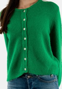Cardigan lavorato a maglia verde con scollatura rotonda, dotato di sei bottoni bianchi, tessuto strutturato e vestibilità rilassata, abbinato a jeans blu.