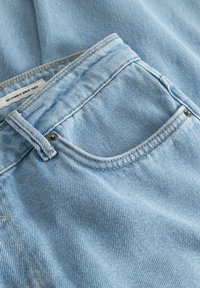 Lyseblå denimjeans brettet, med frontlomme, beltehempe og metallnagler, med synlig tekstur i stoffet og en delvis synlig merkeetikett.