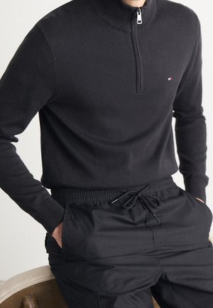 Man wearing a black half-zip sweater with a small logo on the chest, paired with black drawstring pants, hands in pockets.