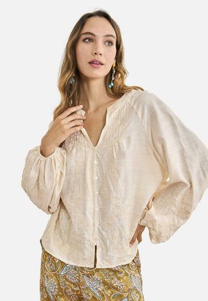 Mujer con una blusa suelta bordada de color crema, con botones y mangas abullonadas, combinada con una falda mostaza con estampado floral.