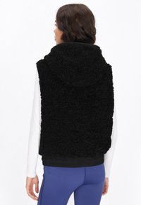 Schwarze Fleece-Weste mit Kapuze, die eine strukturierte Oberfläche hat. Der Saum ist elastisch und die Weste wird über einem weißen Langarmshirt getragen.
