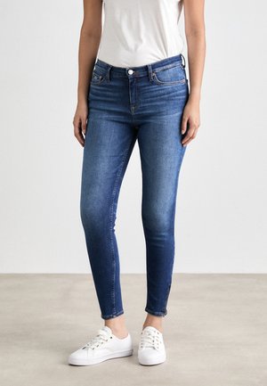 Femme portant un jean bleu ajusté taille moyenne et des baskets blanches, debout sur un sol gris clair contre un mur blanc uni.