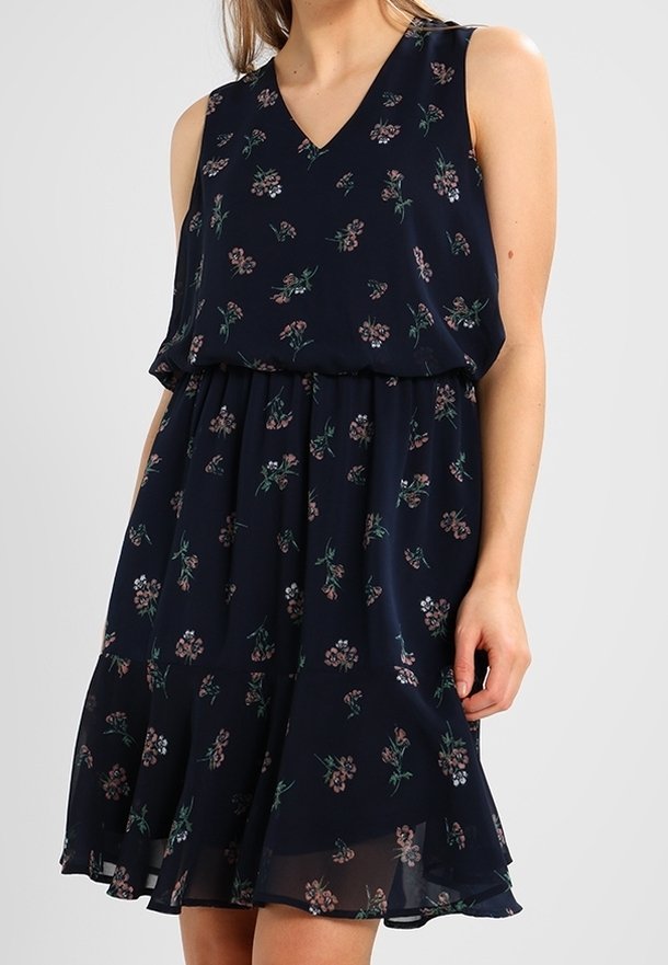 Robe sans manches bleu marine avec motif floral rose, col en V, taille froncée et jupe fluide portée par une personne sur un fond uni.