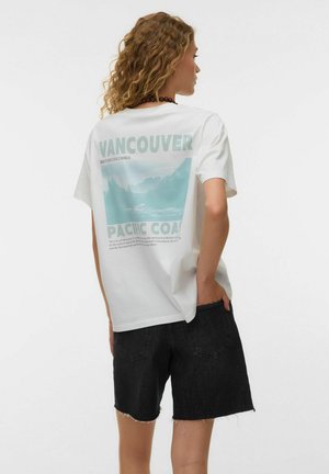 Kvinde med krøllet hår iført hvid Vancouver grafik t-shirt og sorte denim shorts, stående med den ene hånd i lommen, vendt sidelæns.