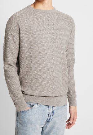 Homme portant un pull en maille texturée gris clair avec col rond et un jean bleu clair, debout devant un fond blanc uni.