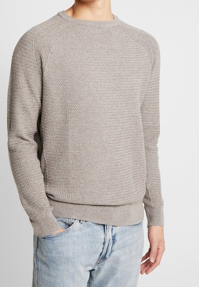 Homme portant un pull en maille texturée gris clair avec col rond et un jean bleu clair, debout devant un fond blanc uni.