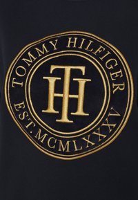 Tessuto nero con un logo circolare ricamato in oro che presenta la scritta "TOMMY HILFIGER" e "EST. MCMLXXXV" in un design ornato.