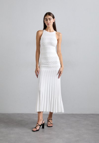 Robe blanche sans manches avec des rayures horizontales texturées, corsage ajusté et jupe plissée évasée. Associée à des sandales à talons avec des lanières noires.