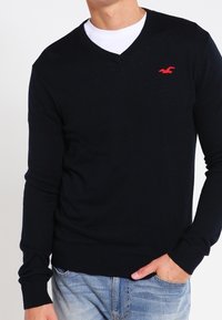 Man som bär en svart V-ringad tröja med en röd logotyp på bröstet över en vit skjorta och ljusblå jeans, med händerna i fickorna.