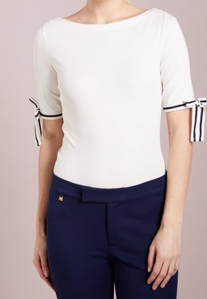 Femme portant un haut blanc ajusté avec des détails de nœud rayé sur les manches, associé à un pantalon taille haute bleu marine.