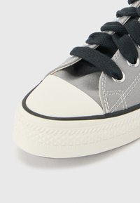 Κοντινό πλάνο γκρι υφασμάτινου αθλητικού παπουτσιού με μαύρα κορδόνια και λευκή ελαστική μύτη και σόλα, σε γκρι φόντο.