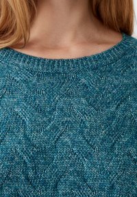 Nahaufnahme einer Frau, die einen blauen, kabelgewebten Pullover mit ribbeligem Rundhalsausschnitt trägt und lange blonde Haare sichtbar sind.
