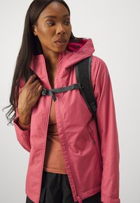 Pink waterproof jacket with a zippered front, adjustable hood, and pockets. Worn over a brown top, paired with a black backpack.