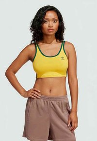 Dzeltena topiņa ar zaļu apdari un mazu logotipu, saskaņota ar bēšiem šortiem. Auduma virsma izskatās gluda ar piegriezumu, kas pielāgots ķermenim.