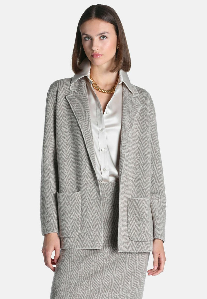Jeune femme aux cheveux bruns et raides porte un blazer gris à chevrons et une jupe sur une blouse en satin blanc avec un collier chaîne doré.