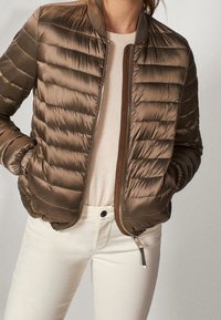 Bruine pufferjack met een gequilte uitstraling, ribbelkraag en een voorkant met rits. Draag het over een lichte trui en crèmekleurige broek.