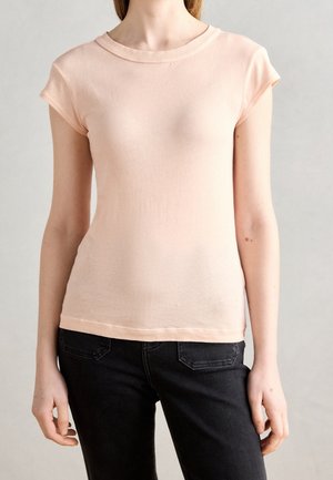 Woman wearing a light pink short-sleeve t-shirt and black jeans, standing with arms relaxed against a neutral gray background.