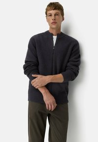 Dunkelgrauer Strickpullover mit einem Reißverschluss, Rippstruktur, runder Kragen und langen Ärmeln, kombiniert mit olivgrünen Hosen.