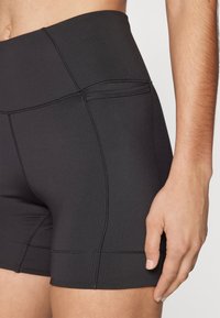 Černé sportovní kraťasy s vysokým pasem, hladkým povrchem a plochými švy pro komfort. Detailní záběr na stranu a ruku.