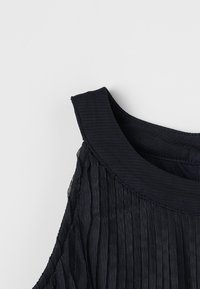 Close-up van een zwart mouwloos jurkje met een textuur, geplooide meshstof en een brede, ronde halslijn.