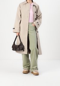 Manteau long beige avec des boutons noirs, haut rose clair, pantalon vert à jambes larges, sac à main marron et chaussures beige avec garniture en fourrure.