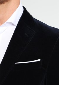 Svart sammetsblazer med snedkragar, som visar en vit fickfäste. Detaljer inkluderar subtil textur och en polerad finish.