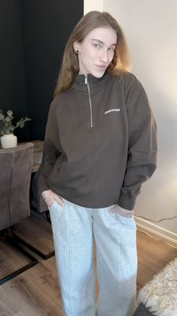 Bruine sweatvest met rits en hoge kraag, voorzien van een wit logo. Gecombineerd met lichtgrijze sweatpants met wijde pijpen, getoond in een goed verlichte binnenomgeving.