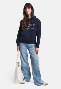 Donkerblauwe hoodie met voorop een logo, kangoeroezak; gecombineerd met lichtblauwe wijde jeans en beige sneakers; houdt een witte tote bag vast.