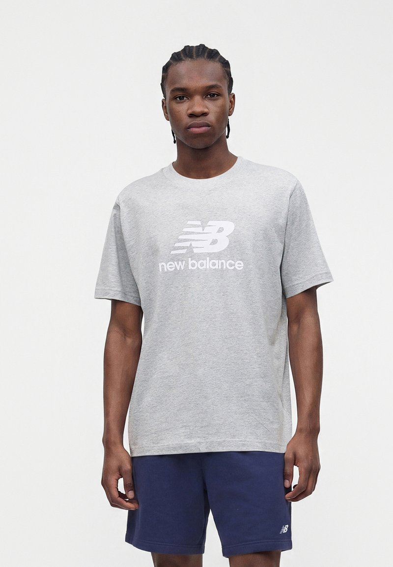 Jonge man met gevlochten haar die een grijs New Balance T-shirt en donkerblauwe korte broek draagt, staand tegen een effen witte achtergrond.