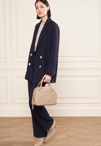 Blazer navy doppiopetto con bottoni bianchi, abbinato a un top beige e pantaloni slim. Tiene una borsa a mano in pelle beige chiaro.