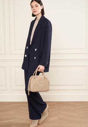 Blazer navy doppiopetto con bottoni bianchi, abbinato a un top beige e pantaloni slim. Tiene una borsa a mano in pelle beige chiaro.