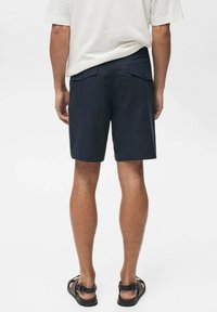 Shorts dark navy cu o textură netedă, două buzunare pe spate și închideri cu nasturi, asortați cu o cămașă lejera de culoare deschisă.
