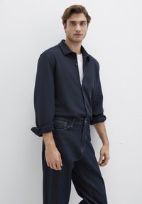 Marineblaues Hemd mit Button-Down-Kragen, hochgekrempelten langen Ärmeln, über einem weißen T-Shirt getragen, kombiniert mit hochtaillierten, dunklen Jeans.