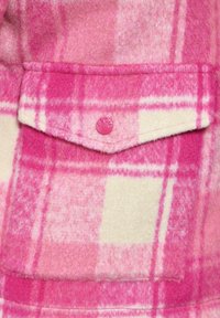 Tissu en polaire à carreaux roses, comprenant des carrés de rose clair et de crème, avec une poche à rabat fermée par un bouton rose. Texture douce dans l'ensemble.