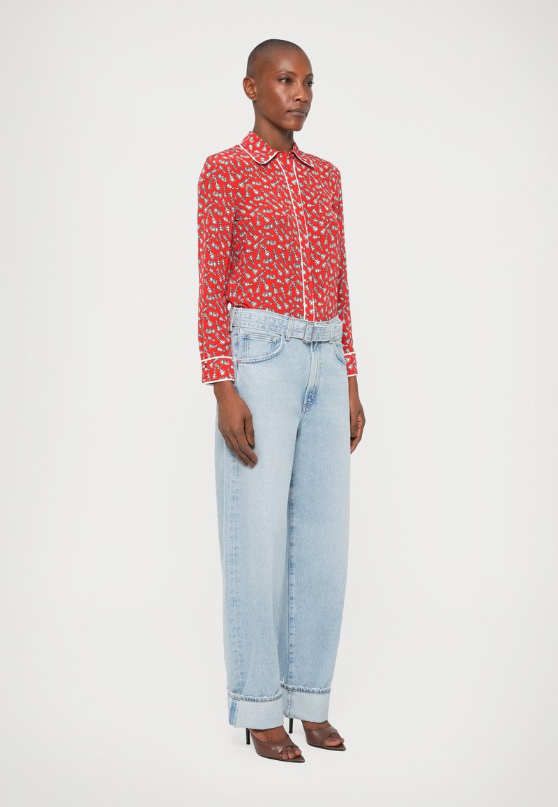 Camisa roja de manga larga con patrones blancos, combinada con jeans de pierna ancha de color azul claro y sandalias de tacón negro. Detalles sencillos en el cuello y los puños.