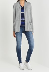 Kvinna iförd en blårandig skjorta, grå öppen cardigan med fickor, blå tighta jeans och vita sneakers, stående mot en vit bakgrund.