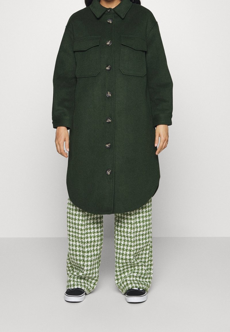 Green wool blend coat with a collared neck, two chest pockets, and row of buttons. Paired with green houndstooth patterned pants and dark sneakers.