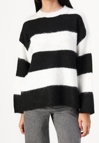 Femme portant un pull duveteux à larges rayures horizontales noires et blanches avec des manches longues et un jean gris clair.