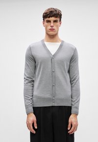 Siva pletena jopica z V-izrezom, rebrastimi rokavi in robom, ki ima šest gumbov spredaj in gladko teksturo. Enostavna ozadje.