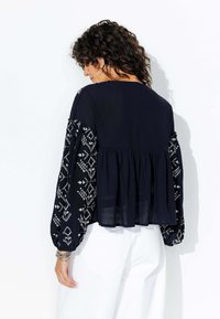 Blouse navy avec de longues manches fluides ornées de motifs floraux brodés en blanc. Coupe évasée avec une taille froncée. Tissu léger.