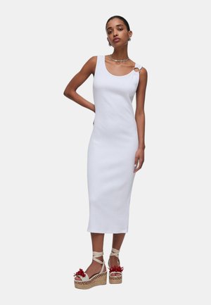 Donna che indossa un abito midi bianco senza maniche con dettaglio ad anello sulla spalla e sandali beige con zeppa e dettagli di fiori rossi.