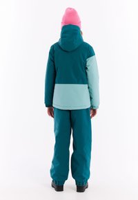 Türkis und hellblaue isolierte Jacke mit Kapuze, kombiniert mit passenden türkisfarbenen Hosen und schwarzen Stiefeln, von hinten betrachtet. Pinkfarbener Beanie getragen.