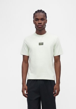 Uomo con capelli trecciati che indossa una maglietta Nike chiara e pantaloni neri, in piedi davanti a uno sfondo bianco semplice.