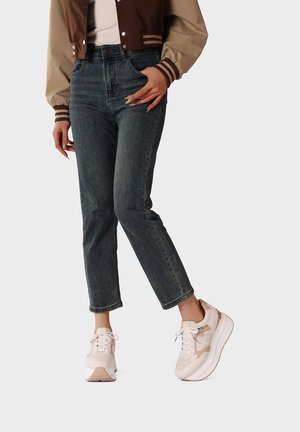 Persona che indossa jeans blu scuro, giacca bomber beige chiaro e marrone, e sneakers beige con suola platform e lacci bianchi, in piedi su sfondo chiaro.