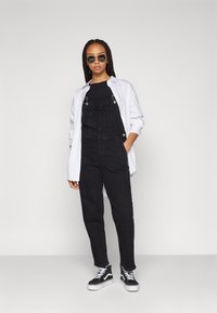 Čierne denimmové overaly so striebornými komponentmi, nosené cez čierny top, skombinované s bielou košeľou a čierno-bielymi teniskami s platformou.