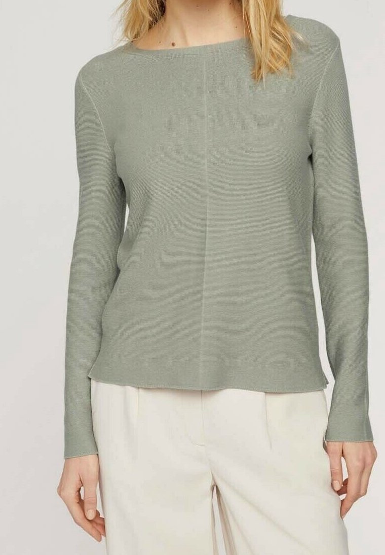 Femme portant un haut en maille vert clair à manches longues avec un col bateau et un pantalon taille haute écru sur un fond uni.