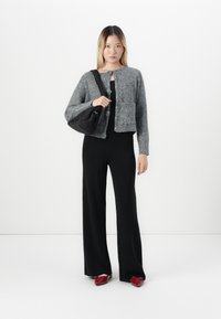 Woman wearing a grey knit cardigan, black wide-leg pants, red shoes, and carrying a black shoulder bag on a white background.
