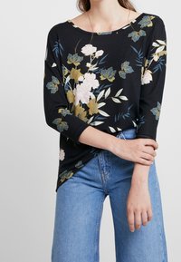 Top floreale nero con maniche a tre quarti, caratterizzato da motivi floreali rosa e verdi, abbinato a jeans blu a vita alta. Texture liscia.