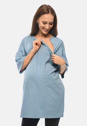 Pregnant woman in a loose light blue top unzipping the front, smiling and looking down against a plain light background.